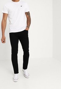 Vit t-shirt med rund halsringning och liten svart logotyp, kombinerad med svarta slim-fit byxor och vita sneakers med röda detaljer.