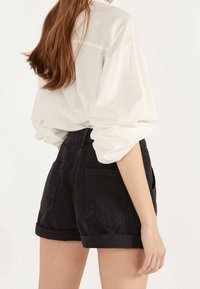 Camisa blanca de manga larga con mangas abullonadas, combinada con pantalones cortos de mezclilla negros con dobladillos enrollados y bolsillos traseros. Diseño simple y moderno.