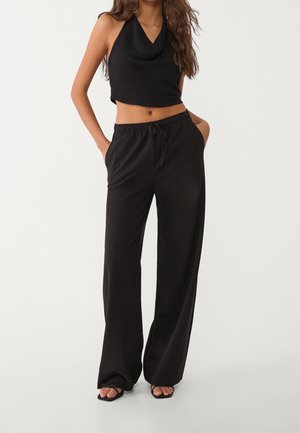 Mujer con top negro sin mangas y corto, y pantalones negros de pierna ancha con cordón en la cintura, de pie con las manos en los bolsillos.