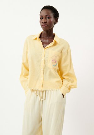 Chemise en lin jaune à boutons avec une poche présentant un motif brodé. Associée à un pantalon à taille élastique à rayures claires.