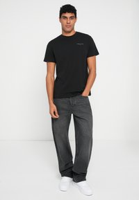 Camiseta de algodón negra de manga corta con un pequeño logo. Combinada con jeans gris oscuro y zapatillas blancas, de pie frente a un fondo liso.