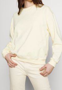 Jasnożółta bluza z okrągłym dekoltem, o luźnym kroju i ściągaczach na rękawach, zestawiona z kremowymi spodniami w paski. Miękka, gładka faktura.
