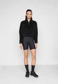 Pull en polaire noir zippé avec un col haut, associé à un short de cyclisme noir à motifs, des chaussettes noires et des chaussures de sport.