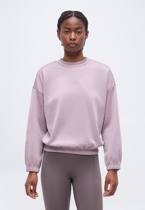 Kobieta z warkoczami, ubrana w jasnofioletową bluzę z długim rękawem i dopasowane szare legginsy, stojąca na tle jednolitego białego tła.