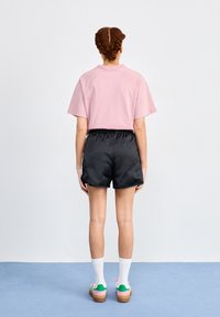 Pink short-sleeve shirt, black nylon shorts, white crew socks, and multicolored sneakers. Model viewed from behind on a light blue floor.