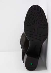 Semelle d'une botte noire avec un motif d'arbre en relief, petit détail hexagonal vert près du talon, inclinée sur un fond blanc.