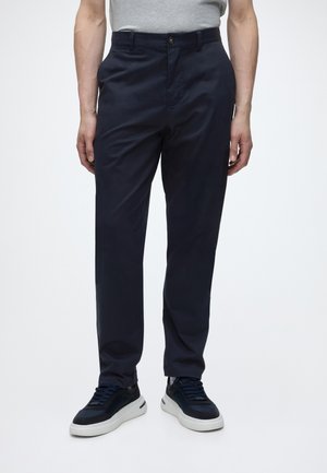 Mann trägt dunkelblaue, gerade geschnittene Hose mit schwarzen und weißen Sneakers, steht vor einem einfarbigen hellen Hintergrund.