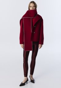 Červený vlněný kabát s dvojitým zapínáním a odpovídajícím dlouhým šálkem, spárovaný s lesklými burgundskými legínami a černými lodičkami se špičatou špičkou.
