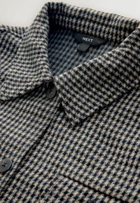 Button-up-Hemd im karierten Muster, das Grautöne, Beige und Marineblau umfasst. Das Material wirkt strukturiert mit einem ausgeprägten Kragen und einem einzelnen Knopf.