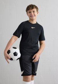 Poiss naeratab, kandes musta Nike jalgpalliülikonda, hoides käes musta-valget jalgpalli, taustaks helehall sein.