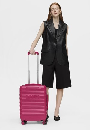 Mujer con cabello largo y rojo, vestida con un blazer sin mangas negro y pantalones cortos, sosteniendo una maleta rosa con ruedas con la palabra "TOUS" en relieve.