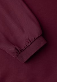 Gros plan sur un sweat-shirt bordeaux présentant une manche froncée et un poignet côtelé, fabriqué en tissu doux à la texture lisse.