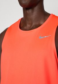 Nærbilde av en person som har på seg en lys oransje ermeløs treningsgenser med en sølv Nike swoosh-logo på brystet.