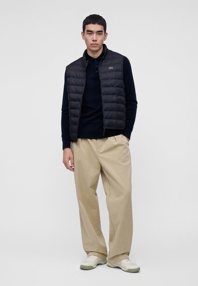 Jeune homme portant une veste sans manches matelassée noire, une chemise noire à manches longues, un pantalon large beige et des baskets blanches, debout devant un fond uni.