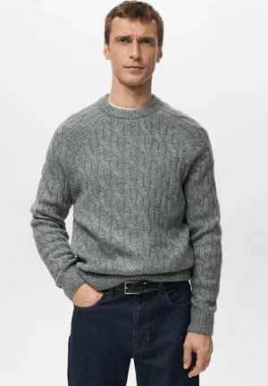 Suéter gris de punto con patrón de cables, cuello redondo y puños acanalados, combinado con vaqueros de mezclilla oscuros.