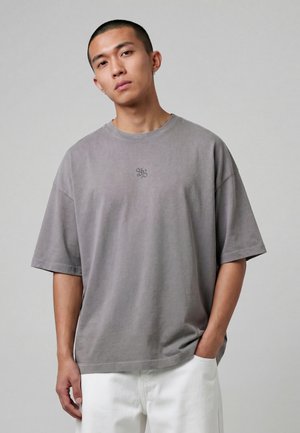 Ung mann med kort hår som har på seg en løs grå T-skjorte med et lite emblem og hvite bukser, stående mot en ensfarget grå bakgrunn.