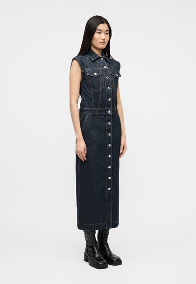 Γυναίκα που φοράει αμάνικο σκούρο τζιν φόρεμα με κουμπιά και μαύρες πλατφόρμες, στέκεται μπροστά σε απλό λευκό φόντο.