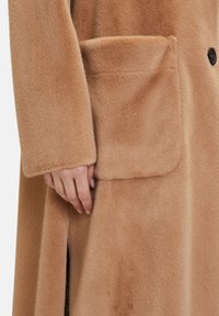 Cappotto in pelliccia beige con una consistenza morbida, dotato di una grande tasca applicata e un singolo bottone nero, mostrato da vicino su una mano.