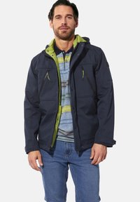 Navy jacket with a green hood, featuring zippered chest pockets and snap buttons. Worn over a striped polo shirt and denim jeans.
