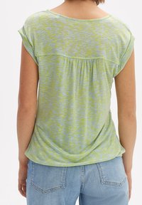 Žena v krátkém rukávu, lehce zeleném vzorovaném topu, kombinovaném s světle modrými džíny, zobrazená zezadu na bílém pozadí.