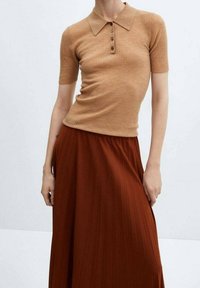 Mujer vistiendo una camiseta polo de manga corta acanalada en color camel y una falda plisada de tiro alto en color óxido frente a un fondo liso.