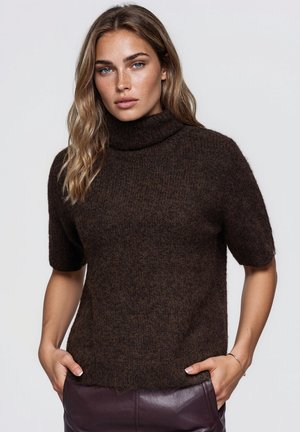 Une femme pose devant un fond uni, vêtue d’un pull à col roulé marron foncé à manches courtes et d’un pantalon en cuir bordeaux.