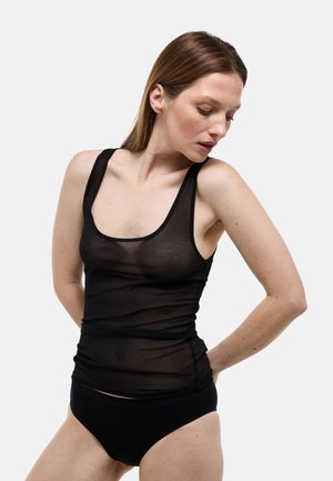 Femme portant un haut noir transparent sans manches et une culotte noire, légèrement de profil avec les mains derrière le dos sur un fond uni.