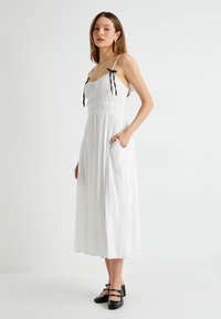 Witte, mouwloze midi-jurk met een gesmokte borstpartij, verstelbare zwarte linten en zijzakken. Model draagt zwarte blokhak schoenen.