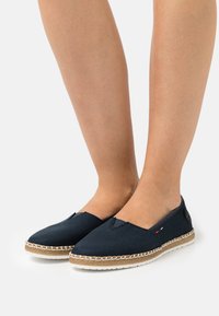Blue canvas slip-on shoes with a rounded toe, woven jute midsole, and contrasting white stitching along the edge.