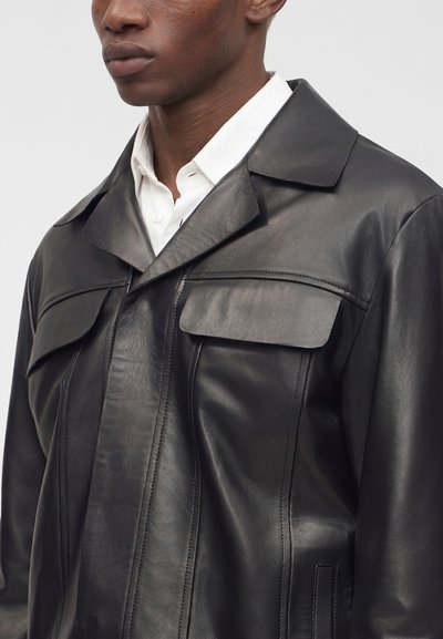 Homme portant une veste en cuir noire par-dessus une chemise blanche boutonnée, montrant le haut du torse et les détails du col.