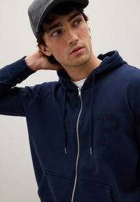 Felpa con zip navy e logo "P" testurizzato, cappuccio con lacci e tasche frontali. Il modello indossa un berretto grigio, su uno sfondo neutro.