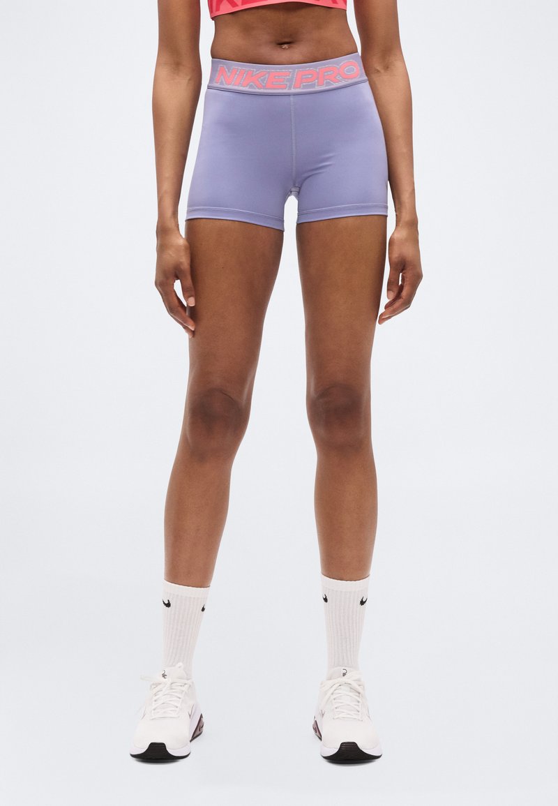 Μωβ σορτς Nike Pro με αντίθετη ροζ ζώνη μέσης, διαθέτουν εφαρμοστή σχεδίαση και λεία υφή. Φοριούνται με λευκές κάλτσες και παπούτσια.