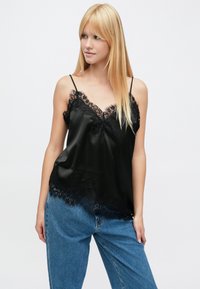 Zwarte satijnen cami met kanten bies langs de halslijn en zoom, voorzien van dunne verstelbare bandjes en een gladde textuur. Gecombineerd met blauwe denim.