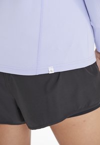 Helllila Sportoberteil mit einem Etikett und schwarzen Shorts, die eine glatte Textur und eine lockere Passform aufweisen.