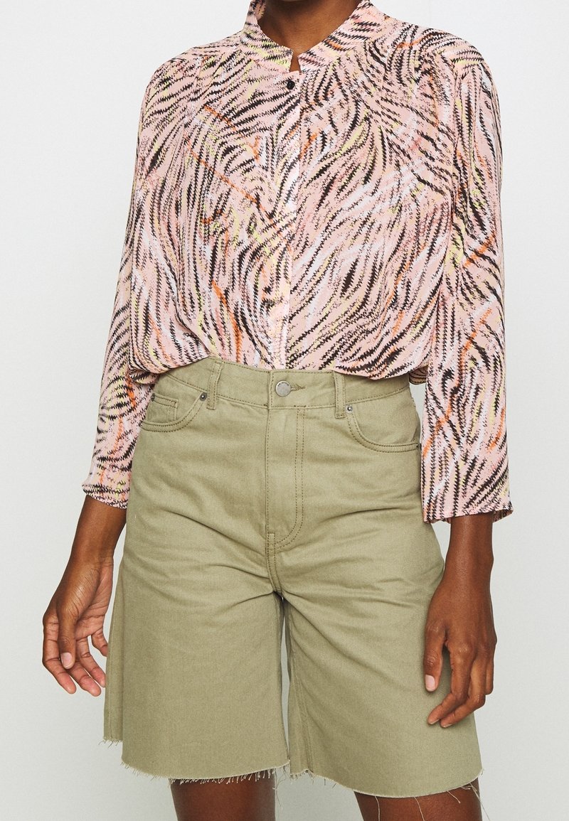 Personne portant une chemise rose à motifs à manches longues, rentrée dans un short taille haute, vert olive clair, longueur genou, avec un ourlet brut.