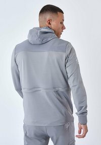 Felpa sportiva grigia con finitura testurizzata, maniche lunghe, cappuccio regolabile e dettaglio della cucitura centrale, abbinata a pantaloni coordinati.