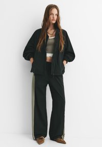 Jachetă neagră ușoară cu manșete elastice, asortată cu pantaloni largi negri cu dungi aurii pe lateral și un top scurt gri. Pantofi maro.