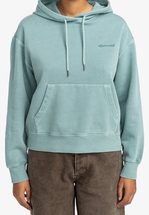 Person iført en lyseblå hættetrøje med forlomme og "element"-logo, kombineret med brune bukser.