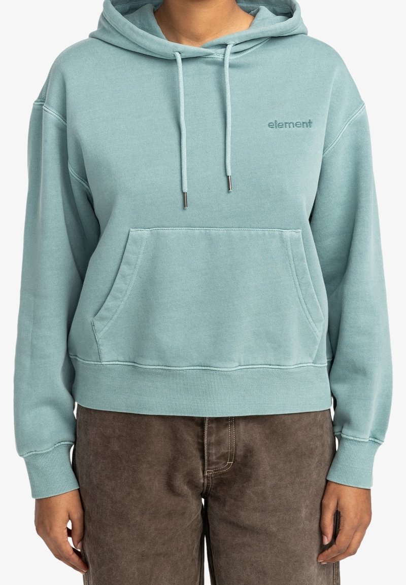 Personne portant un sweat à capuche bleu clair avec poche avant et logo « element », associé à un pantalon marron.