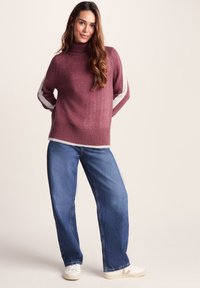 Maroon sweater with a high neck, textured body, and white stripes on sleeves. Paired with loose blue jeans and white shoes.