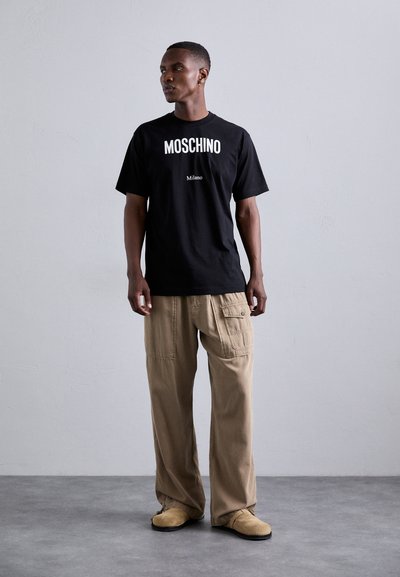 Mustmust T-särk musta värviga, millel on valged tekstid "MOSCHINO" ja "Milano". Beežid cargo püksid taskutega. Pruunid libisemiskindlad kingad. Minimalistlik taust.