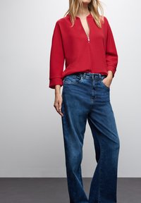 Top rosso con zip, scollatura a V e maniche larghe, abbinato a jeans scuri a vita alta, a vestibilità rilassata, con una leggera scoloritura.