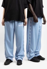 Light blue track pants made of smooth fabric, featuring side stripes with logo patterns and black sneakers. Two models standing side by side.