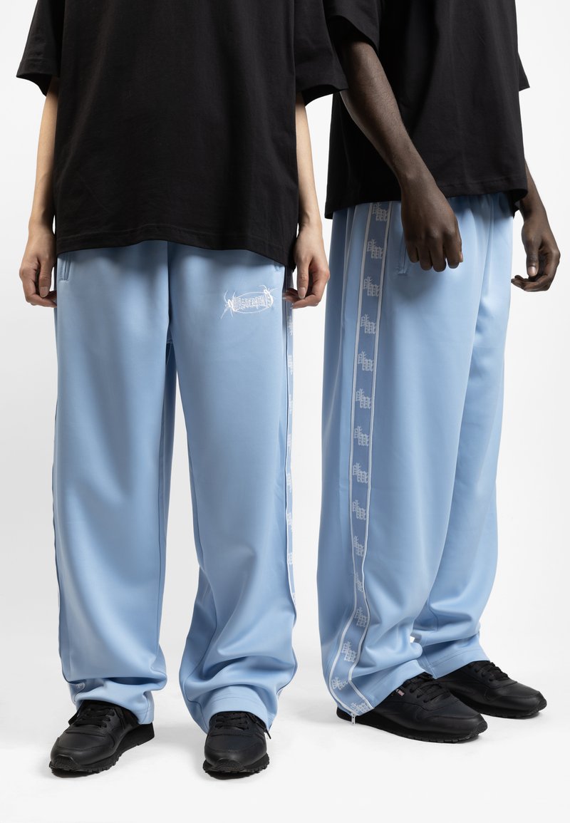 Light blue track pants made of smooth fabric, featuring side stripes with logo patterns and black sneakers. Two models standing side by side.