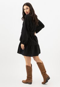 Robe noire à manches longues avec taille élastique et ourlet à volants, portée avec des bottes en suede marron à hauteur de genou avec des bords inclinés.