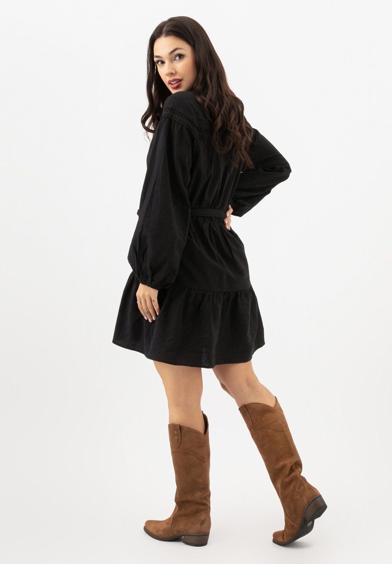 Robe noire à manches longues avec taille élastique et ourlet à volants, portée avec des bottes en suede marron à hauteur de genou avec des bords inclinés.
