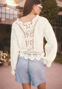 White embroidered blouse with lace detailing, open back, and long sleeves, paired with light blue shorts. Textured fabric and lace accents visible.