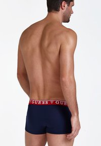 Boxeri pentru bărbați de culoare albastru închis, cu un elastic la talie cu logo-ul roșu "GUESS", având o textură netedă și o potrivire strânsă, prezentat din spate.