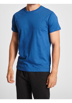 Homme portant un t-shirt bleu uni à manches courtes et un pantalon noir, debout devant un fond gris clair uni.