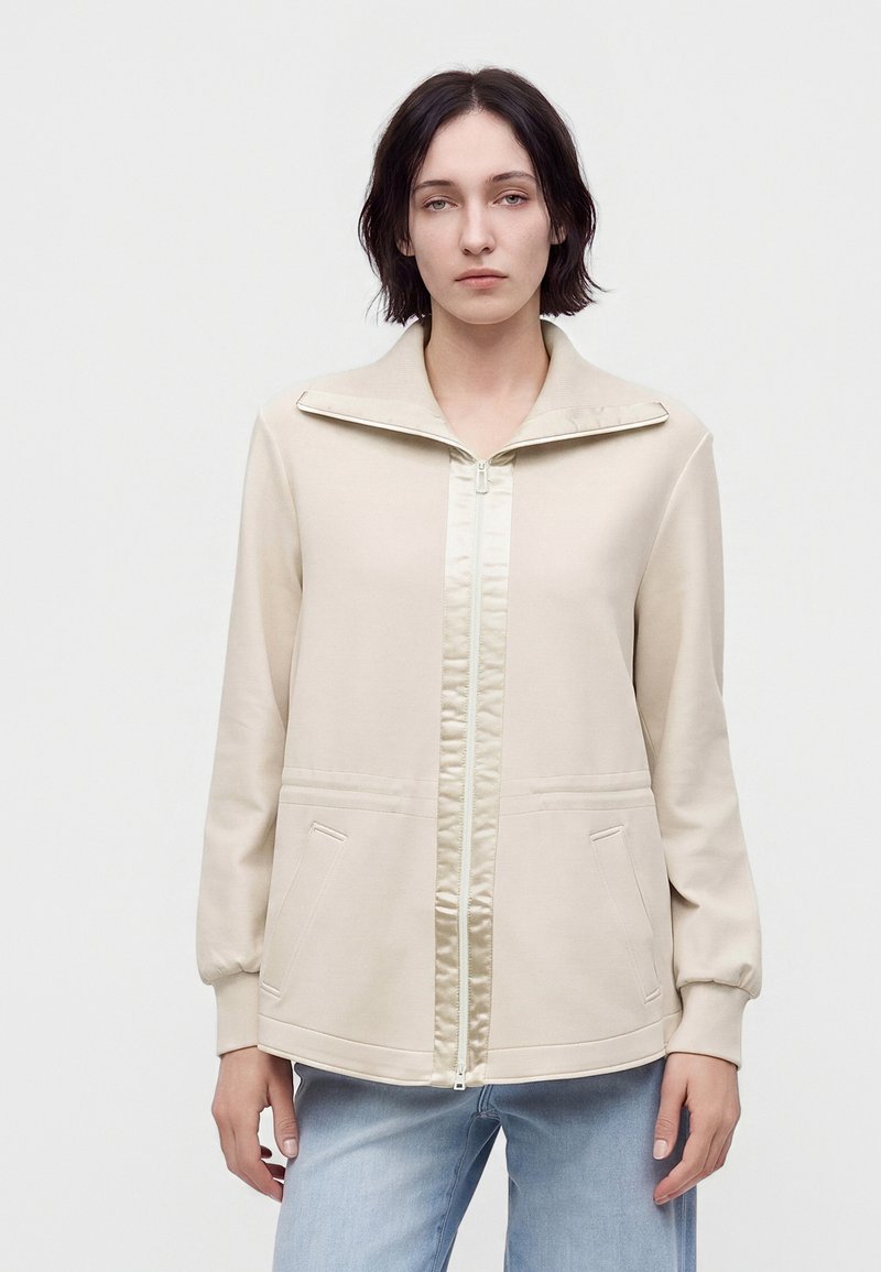 Femme aux cheveux courts et foncés, portant une veste beige zippée et un jean bleu clair, debout devant un fond clair uni.